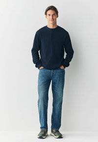 Młody mężczyzna stoi przy białej ścianie, ubrany w granatową bluzkę henley z wafelkiem, niebieskie dżinsy i wielokolorowe sneakersy, z rękami w kieszeniach.