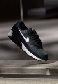 Svart og mørkegrønn Nike Air Max-sneaker med hvit swoosh og såle, som svever over et betongtrinn i en svakt opplyst setting.