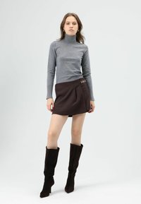 Grijze coltrui gecombineerd met een bruine minirok met een zijden kettingdetail. Model draagt hoge, donkerbruine suède laarzen.