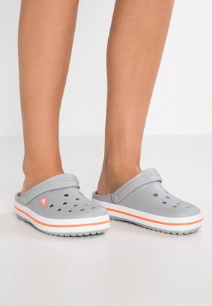 Jambes portant des sabots gris sans lacets avec des trous de ventilation et des semelles rayées orange et blanc sur un sol blanc lisse.