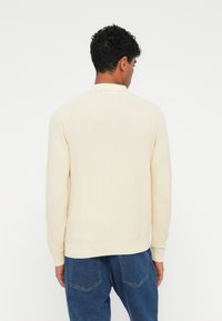 Maglione a maglia beige chiaro con un motivo testurizzato e polsini a coste. Design semplice, collo rotondo, indossato sopra pantaloni in denim blu.