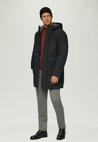 Veste extérieure gris foncé avec capuche, dotée de deux poches avant, fermeture éclair, portée sur un haut bordeaux et un pantalon gris clair, associée à des bottes noires.
