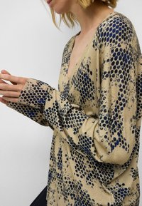 Beige v-hals trui met blauw stippenpatroon, lange mouwen met wijde cuffs, zachte gebreide textuur die de rekbaarheid en val van de stof benadrukt.