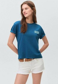 Modré bavlněné tričko s krátkým rukávem a ohrnutými rukávy, na přední straně s bílým nápisem "Chasing Sunsets". V kombinaci s bílými šortkami a páskem.