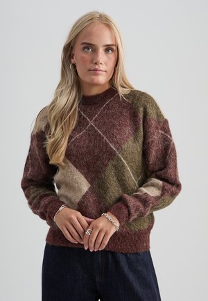 Blonde vrouw die een pluizige, meerkleurige bruine, olijfkleurige en beige trui met geometrisch patroon draagt, met zilveren armbanden en ringen.