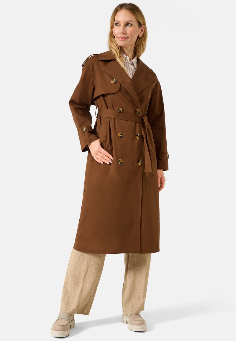Cappotto lunghezza trench marrone con chiusura doppiopetto, grandi bottoni, vita con cintura e ampio colletto. Indossato sopra pantaloni di lino chiari e scarpe.