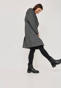 Manteau texturé gris avec une coupe ample, associé à un jean noir slim et des bottines noires à talons avec des détails de boucle sur le côté.