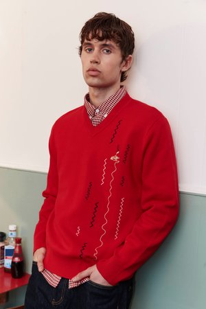 Giovane uomo con capelli castani corti che si appoggia al muro, indossa un maglione rosso con ricami a zigzag neri e bianchi e una camicia a quadri.