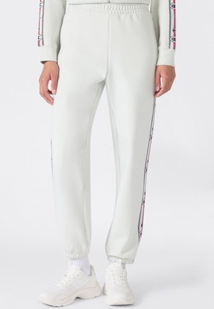 Pantalones deportivos blancos con puños elásticos y rayas laterales con un diseño de logo repetido, usados con zapatillas blancas y calcetines blancos.