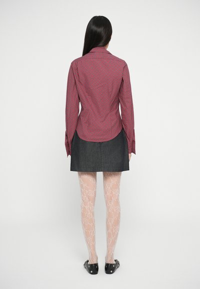 Camisa de botão com padrão vermelho e azul, de mangas compridas, combinada com uma saia de ganga escura, meias-calças com padrão de renda e sapatos planos pretos.