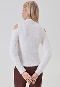 Pull blanc à épaules dénudées, côtelé, avec une texture douce et duveteuse. Comprend des manches ajustées et un petit logo au dos.