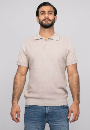 Homme aux cheveux courts et foncés avec barbe, portant un polo en maille beige à manches courtes et un jean bleu, debout devant un fond gris clair uni.