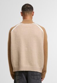 Tansweater in Beige mit hohem Kragen. Weicher Stoff, Design mit fallenden Schultern und geripptem Saum. Weißer Akzent entlang der Schulternaht.
