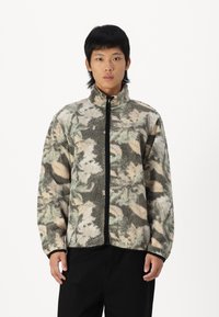 Fleece jacket with a high collar, featuring a floral camouflage pattern in greens, browns, and beige; black zip and cuffs.