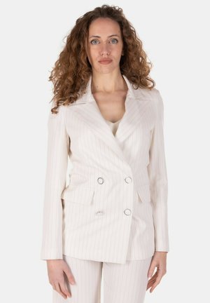Donna con capelli ricci che indossa un blazer doppiopetto a righe bianche e pantaloni abbinati su uno sfondo semplice.