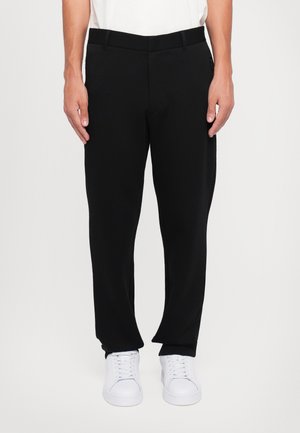 Homme portant un pantalon noir droit, des baskets blanches et une chemise blanche, debout, les mains relâchées le long du corps devant un fond uni.