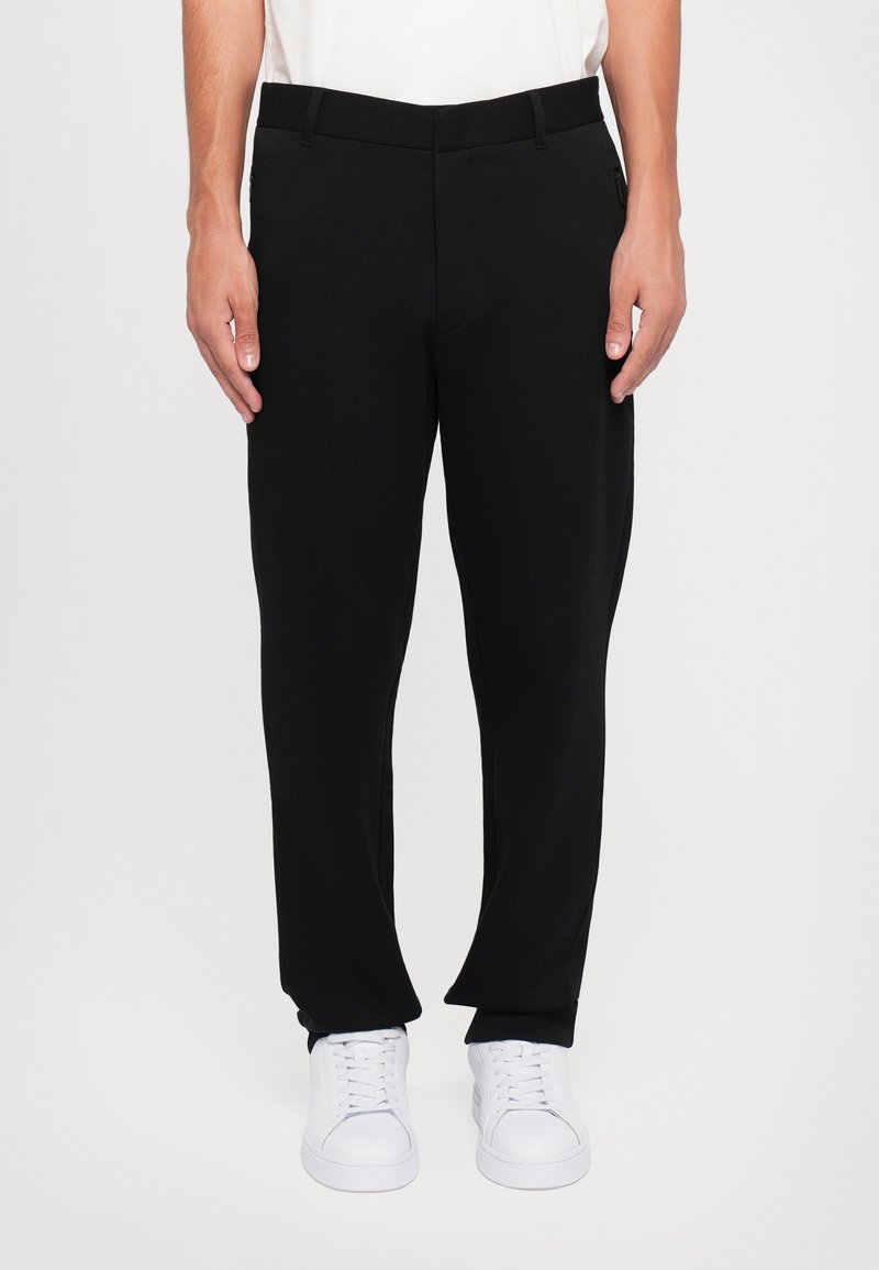 Homme portant un pantalon noir droit, des baskets blanches et une chemise blanche, debout, les mains relâchées le long du corps devant un fond uni.