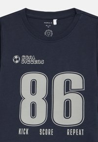 Námorně modré tričko pro batole s velkým potiskem "86", ikonou fotbalového míče a frázemi "Cíle hrabáči", "Kopni", "Skóruj" a "Opakuj" v bílé barvě.