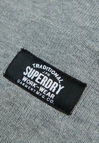 Tissu gris avec une étiquette rectangulaire noire comportant du texte blanc : "TRADITIONAL JPN SUPERDRY WORK-WEAR GARMENT MFG. CO." visible.