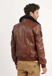 Veste bomber en cuir marron avec un col en shearling noir, des poignets côtelés et une taille côtelée. Elle possède un patch rond sur la manche gauche.