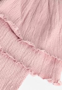 Ljust rosa tyg med fin ribbad textur, med vågiga kanter och mjuka sömmar. Två överlappande lager synliga, vilket framhäver den känsliga designen.