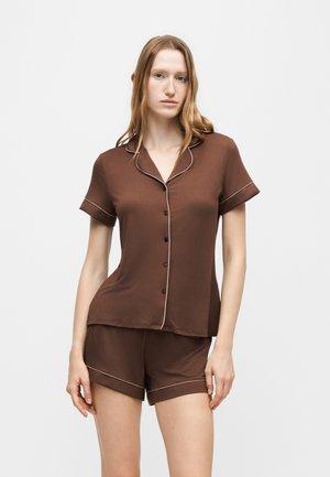 Jeune femme portant une chemise marron à manches courtes boutonnée et un short marron assorti avec passepoil clair sur les bords.