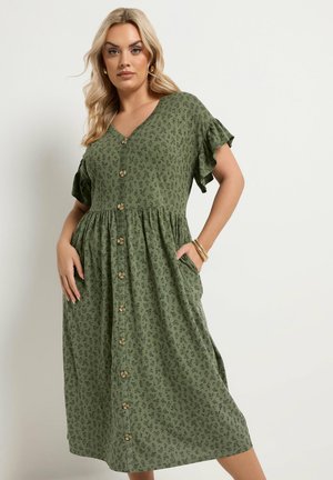 Blondynka w zielonej sukience midi na guziki z krótkimi falowanymi rękawami i kieszeniami, pozująca z ręką w kieszeni na białym tle.