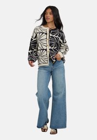 Cardigan en tricot crème et marine avec un grand motif floral, col rond et boutons dorés ; associé à un jean en denim à jambes larges.