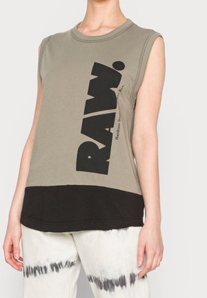 Haut sans manches beige et noir avec un grand texte noir "RAW", porté avec un pantalon blanc avec des rayures tie-dye noires sur les cuisses.