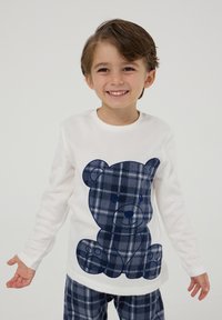 Biała koszula z długim rękawem z dużym, granatowym wzorem w kratę przedstawiającym misia; zestawiona z granatowymi spodniami w kratę. Materiał wydaje się miękki i przytulny.