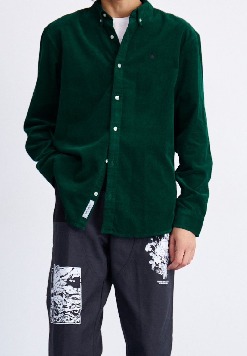 Zielona koszula ze sztruksu z kołnierzem zapinanym na guziki i białymi guzikami, noszona z szarymi spodniami z białym kwiatowym nadrukiem.