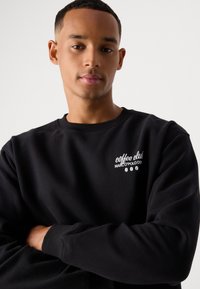 Schwarzer Sweatshirt mit Rundhalsausschnitt und gerippten Bündchen. Mit dem weißen, gestickten Text "coffee club" und "MARCO POLO DE" auf der Brust.