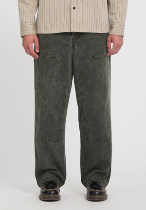 Personne portant un pantalon ample vert foncé avec de grandes poches avant, une chemise rayée beige et des chaussures noires à semelles épaisses.