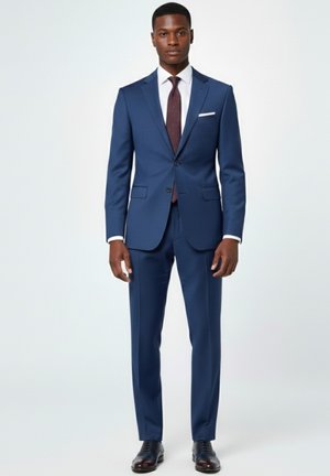 Hombre con traje azul marino a medida, camisa blanca, corbata burdeos con patrón y zapatos de vestir negros, de pie frente a un fondo claro y liso.