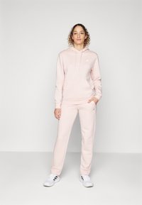 Hoodie rosa claro com um bolso frontal e cordão, combinado com calças de estilo jogging em coordenação. Apresenta um pequeno logotipo branco no peito. Ténis brancos.