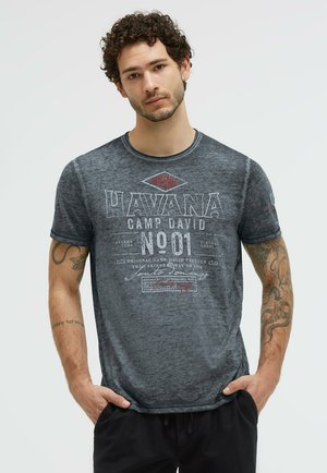 Young man with curly hair and tattoos wearing a faded dark gray t-shirt with "HAVANA CAMP DAVID NO 01" text, standing with hands in pockets.