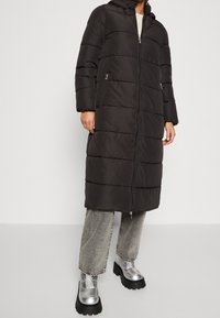 Black puffer coat with a hood, featuring horizontal quilted sections, front zip closure, and side pockets. Worn over gray jeans and silver boots.