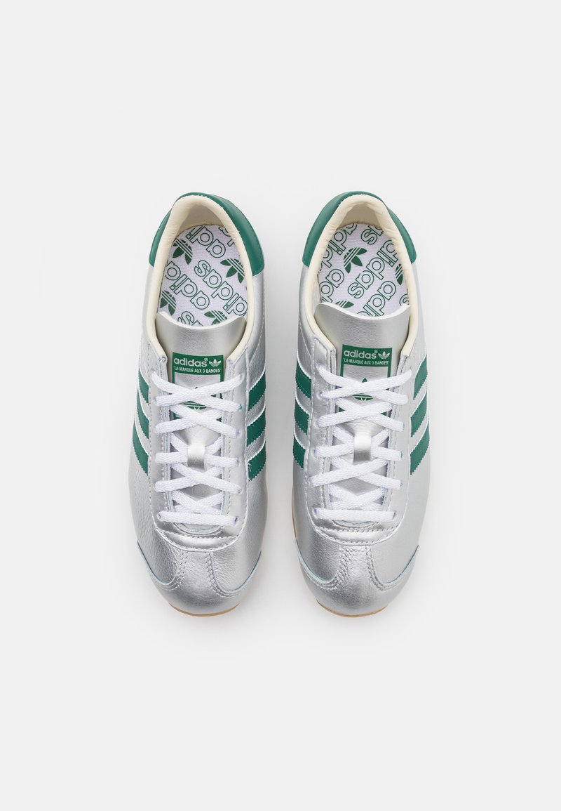 Srebrne buty sportowe z białymi sznurówkami, zielonymi paskami i wzorzystą wkładką z logo Adidas. Gładka tekstura i zaokrąglony kształt.