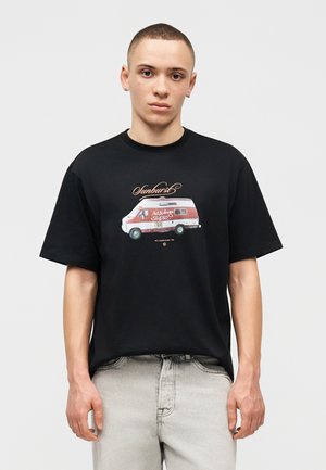 Νεαρός με κοντά μαλλιά φοράει μαύρο T-shirt με κόκκινο και λευκό γραφικό βαν και τη λέξη "Sunburst", συνδυασμένο με ανοιχτό γκρι τζιν.