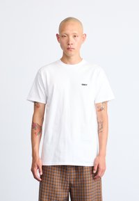 White cotton t-shirt with a round neckline and short sleeves, featuring a small black "OBEY" logo on the left chest. Patterned brown checkered pants.