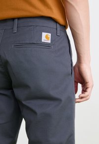 Graue Baumwollhosen verfügen über eine Rückentasche mit einem Logo-Tag. Der Bund ist eng anliegend, und der Stoff hat eine glatte, haltbare Textur.