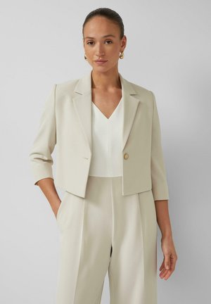 Frau trägt beige gekürzten Blazer mit einem Knopf über weißem Top und passenden hoch taillierten weiten Hosen, steht mit Hand in der Tasche.