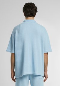 Light blue oversized shirt made of textured fabric with short sleeves, a collar, and a straight hem. Design features subtle vertical ribbing.