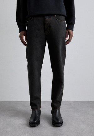 Person iført sorte jeans med lige ben og sorte læder ankelstøvler, stående på en grå gulv mod en lysegrå væg.