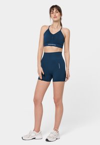 Laivastonsininen urheiluliivi ja siihen sopivat korkeavyötäröiset shortsit joustavasta kankaasta. Liivissä on rypytetty etuosa; shortseissa on logo sivussa.