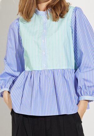 Femme portant un chemisier ample à manches longues avec des rayures blanches verticales sur des panneaux bleu clair et vert menthe, associé à un pantalon noir.