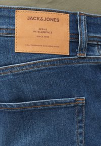 Jeans in denim con una tonalità blu scuro, cuciture arancioni e una toppa rettangolare in pelle con il nome del marchio "JACK & JONES" attaccata alla vita.