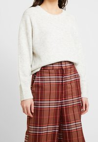 Hellgrauer Strickpullover mit Rundhalsausschnitt und langen Ärmeln, kombiniert mit hochtaillierten, karierten weit geschnittenen Hosen in Rot-, Orange- und Violetttönen.