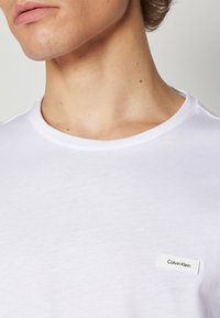 White crew neck T-shirt made of soft cotton; features a small branded tag on the left chest with "Calvin Klein" in black text.