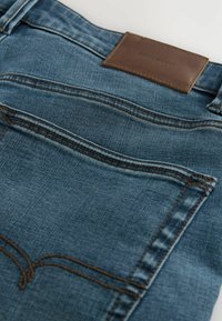 Jean en denim de couleur bleu foncé, avec une patte en cuir marron à la taille et des coutures contrastées sur les poches arrière.